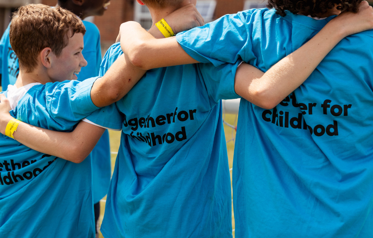 3 young boys with their arms around each other facing away from the camera, the boy on the left is smiling at the other two boys. they are all wearing blue thsirts with 'together for childhood' written on the back in black font. they are in front of a brick building
